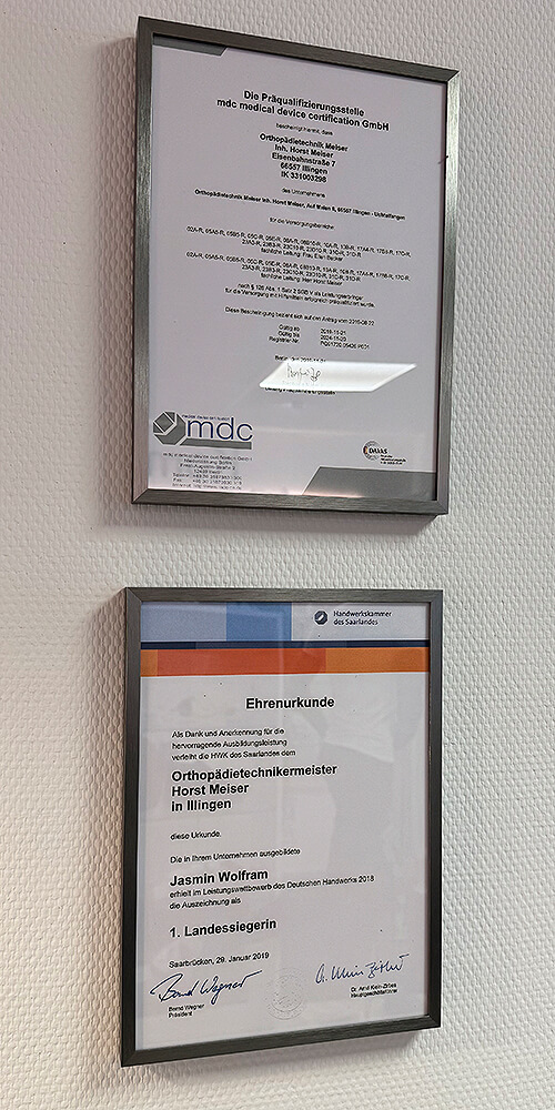 Zwei gerahmte Urkunden und Zertifikate hängen an einer hellen Wand in einem Flur